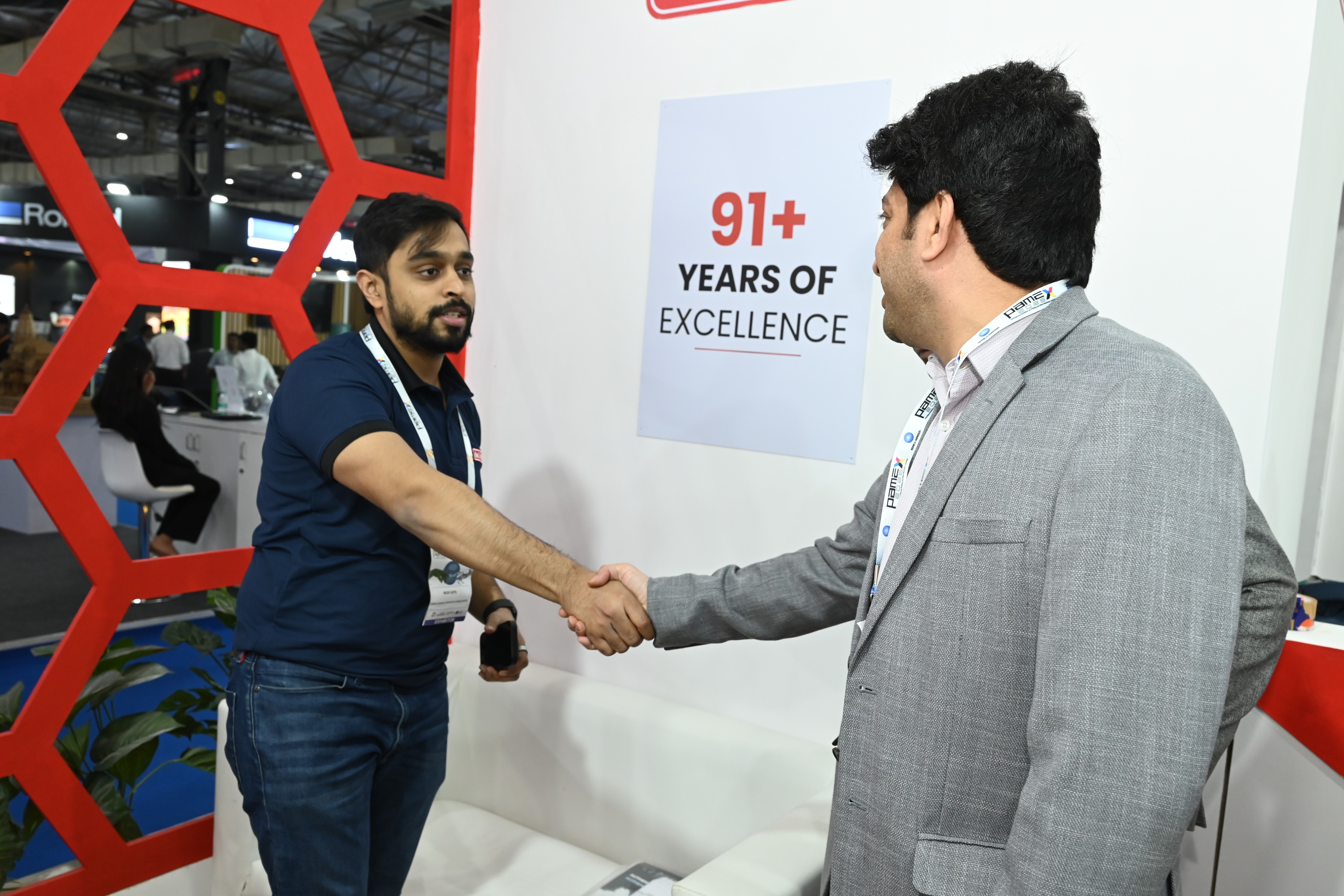 A Desmat team member shaking hands with a visitor beside the 91+ Years of Excellence panel